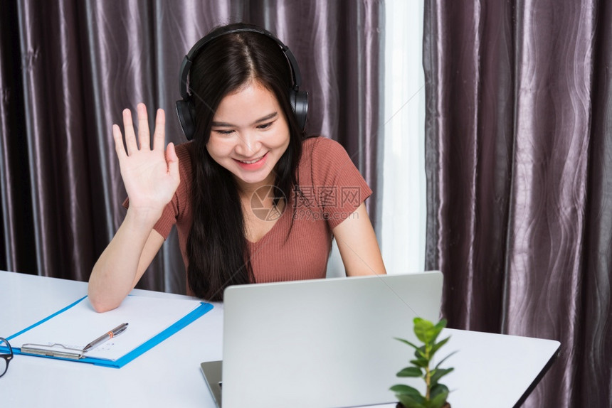 在家工作笑的亚洲快乐商业坐在办公桌工作空间的年轻美女戴着耳机视频电话会议带笔记本电脑举手打招呼在家庭办公室打招呼图片