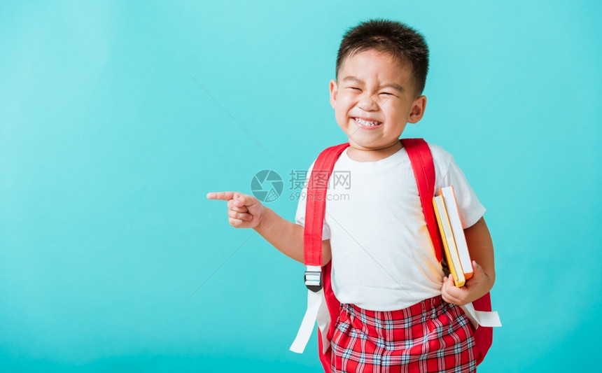 长相快乐的可爱亚洲小孩男笑着拥抱书和指向空间孤立的蓝背景图片