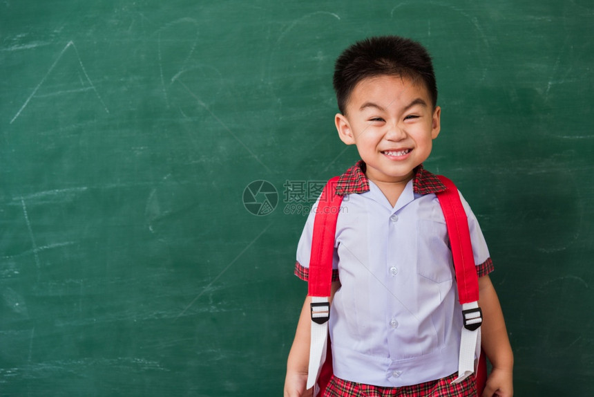 快乐的亚洲可爱小孩男从幼儿园的稚身着校服学生穿拿书包站在绿色学校的黑板上微笑第一次到学校教育概念图片