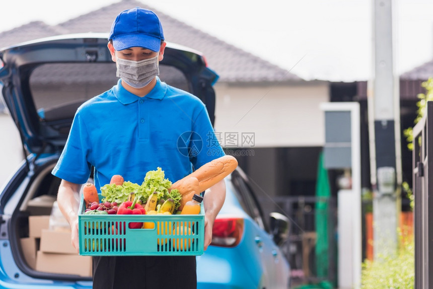 亚洲杂货店送员身穿蓝色制服面罩保护身穿蓝色制服在科罗纳爆发后在门前家的塑料箱中提供新鲜食品蔬菜回到新的正常概念图片