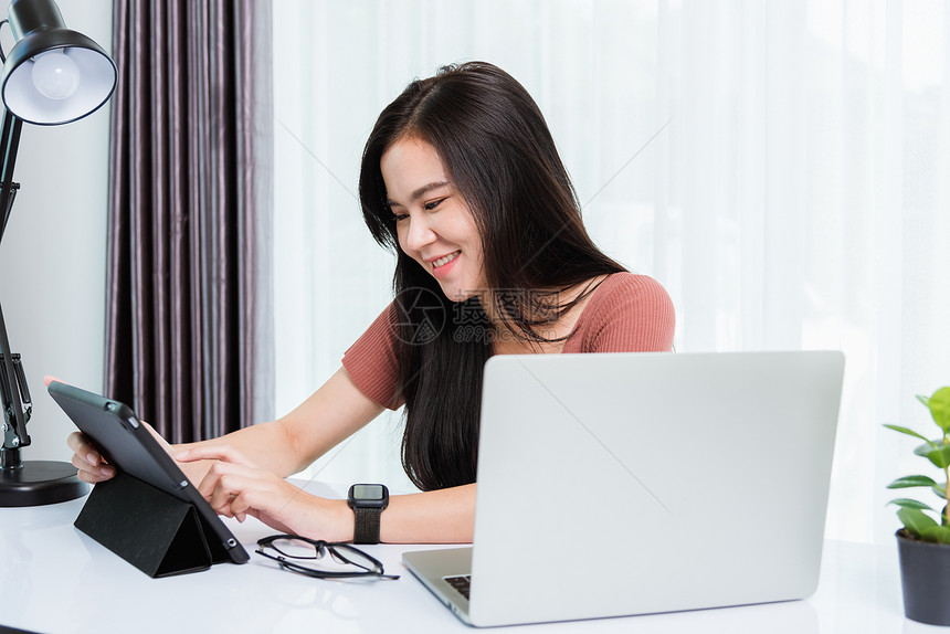 在家工作微笑着快乐的亚洲商界工作坐在办公桌工作空间的年轻美丽女士使用现代智能平板电脑和膝上型计算机技术电脑视频话会议在家庭办公室图片
