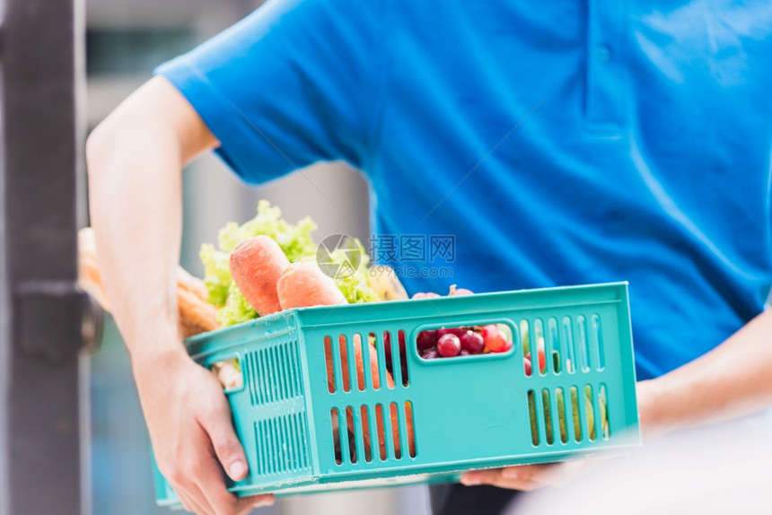 亚洲杂货店送员身穿蓝色制服面罩保护身穿蓝色制服在科罗纳爆发后在门前家的塑料箱中提供新鲜食品蔬菜回到新的正常概念图片