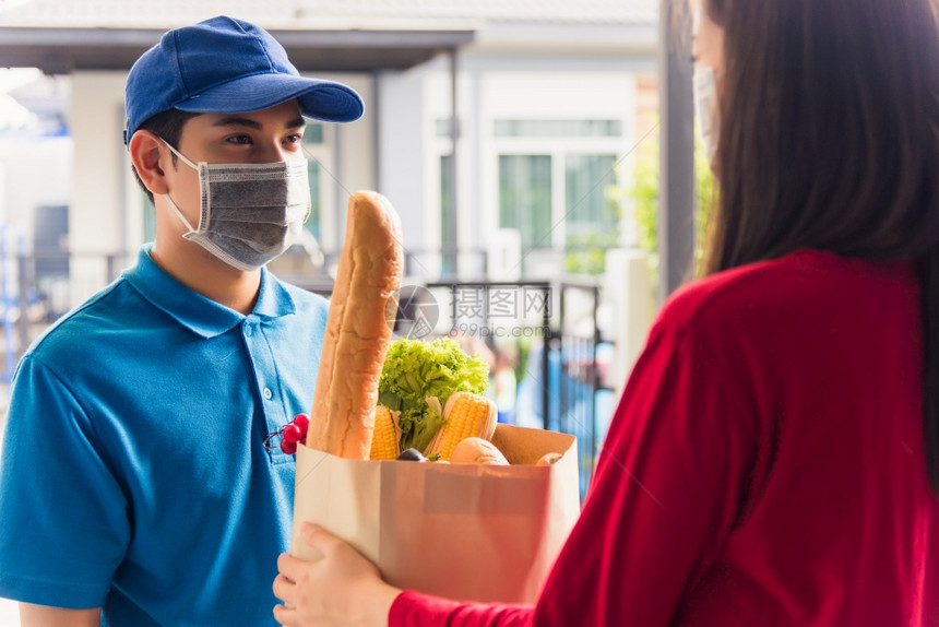 身穿制服的亚洲青年送货员戴保护面罩他制作杂货服务根据流行冠状向接受前门服务的女顾客提供新鲜食品回到的正常概念图片