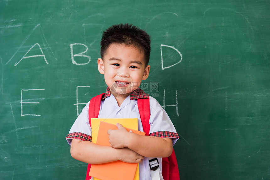 快乐的亚洲可爱小男孩幼儿园穿着校服学生制拿书包和笑在绿色学校黑板上显示手指拇第一次上学图片