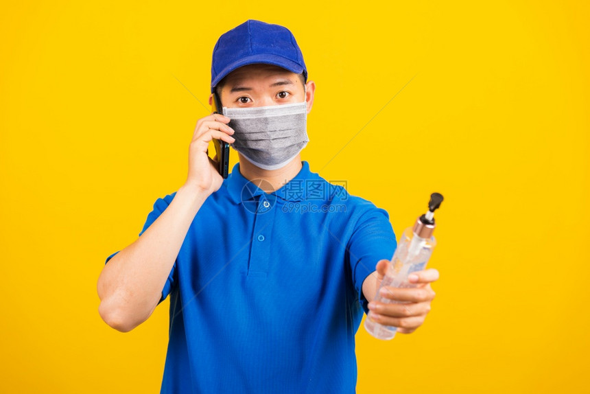亚洲年轻英俊的送货员身着面罩保护持有酒精洗净凝胶移动电话工作室打给顾客拍摄孤立的黄色背景爆发的科罗纳COVID19图片