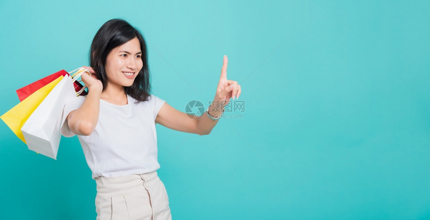 身穿白T恤的亚洲美丽年轻女子站立她手拿着袋持多色摸指的空气在工作室拍摄蓝色背景的照片图片