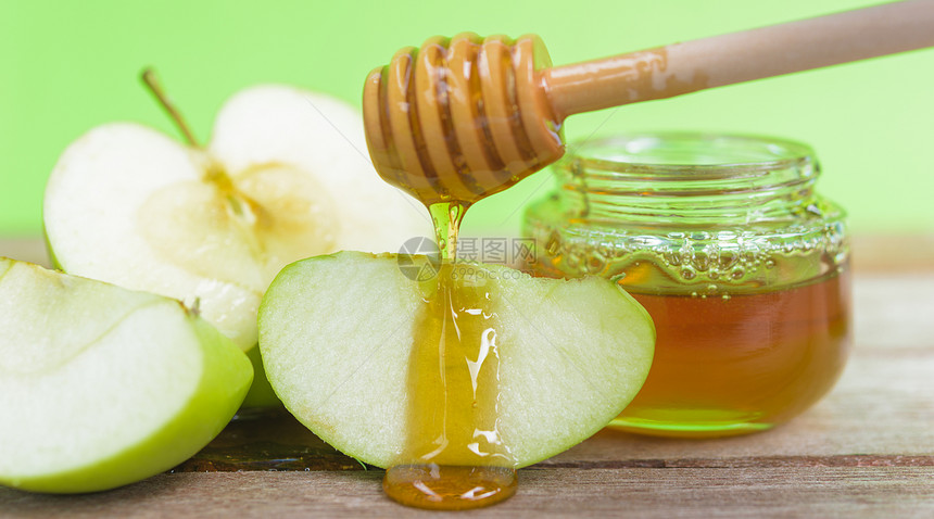 犹太节日概念苹果罗什哈沙纳甜点日照片上的中蜂蜜在一个罐子里蜂蜜在绿苹果上落下被切在绿色背景的木头上图片