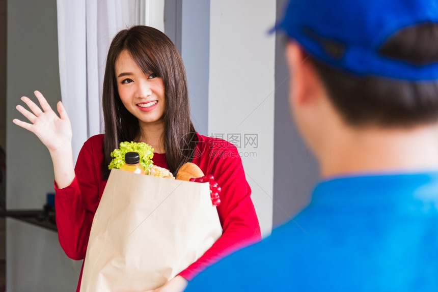 蓝制服亚洲青年送货员制作杂服务在流行冠状后用纸袋向前门女顾客提供新鲜蔬菜水果和食品回到新的正常状态图片