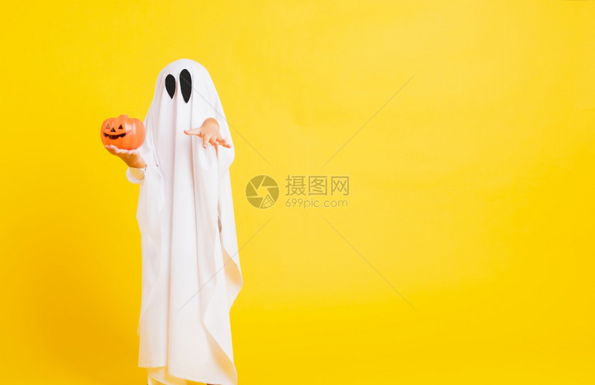 有趣的万圣节基概念可爱的小孩与白色服装扮神圣的灵吓他手握着橙南瓜的鬼摄影棚拍黄色的白背景图片