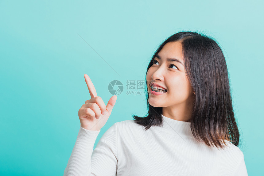 亚洲年轻美丽的女子微笑肖像有牙套尖笑的手指边远离空白复制间以蓝背景医学和牙概念孤立拍摄的影棚图片