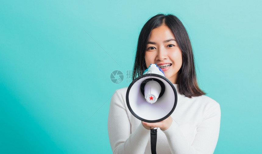 美丽的亚洲女微笑她拿着扩音器做宣布女兴奋的欢快乐宣布好消息孤立在蓝背景上图片