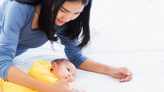 美丽年轻的亚洲母亲用白床上的奶瓶喂养婴儿图片
