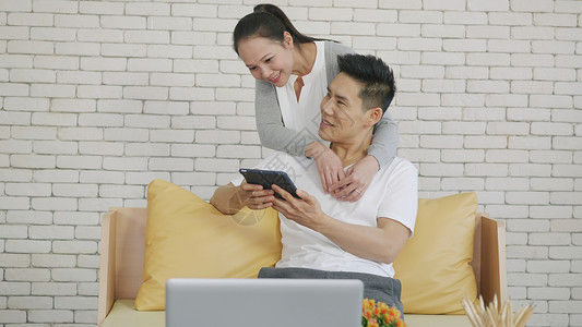 亚洲家庭夫妇妻一起工作使用数字平板电脑幸福的爱侣们笑着在网上购物从家里在客厅看社交媒体背景图片