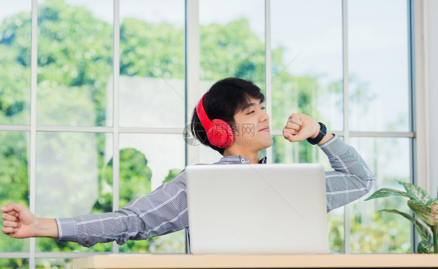 亚洲青年商人在家办公室用红耳机微笑听音乐坐在办公桌上放松穿戴无线头电话的快乐商人享受听音和笔记本电脑的工作图片