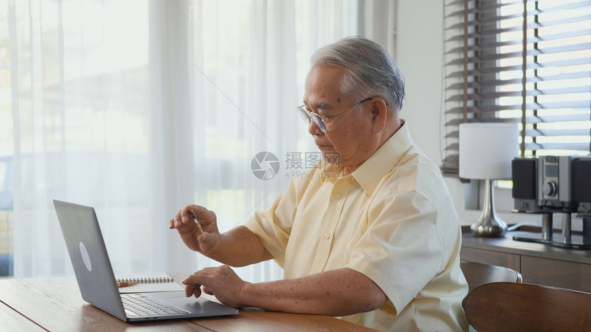 在家客厅用笔记本电脑工作时坐在椅子上工作时戴眼镜的老人快乐老人用电脑退休老人祖父在家工作图片