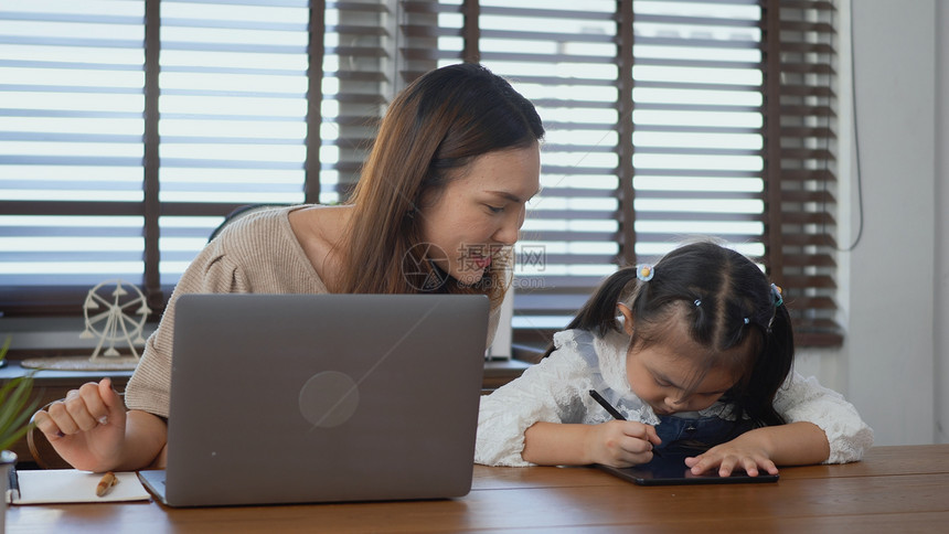 亚洲年轻母亲在家使用笔记本电脑帮助女儿做家庭作业单身母亲自由职业者在家工作帮助其女孩在书桌上做家庭作业图片