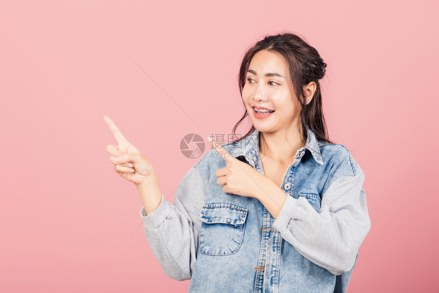 亚洲快乐的肖像美丽可爱的年轻女子穿着戴尼姆站指对展示产品看对面摄影棚拍着粉红背景与复制空间隔绝图片