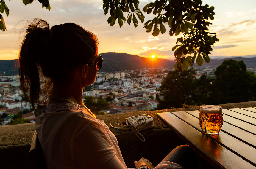 奥地利格拉茨市一位年轻女子坐在那里手里拿着一杯啤酒看着夕阳从Schlossberg山著名旅游景点钟楼的餐厅俯瞰奥地利格拉茨市一位图片