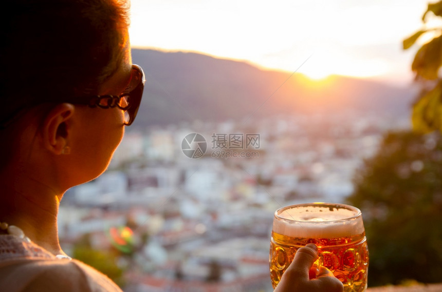 奥地利格拉茨市一位年轻女子坐在那里手里拿着一杯啤酒看着夕阳从Schlossberg山著名旅游景点钟楼的餐厅俯瞰奥地利格拉茨市一位图片