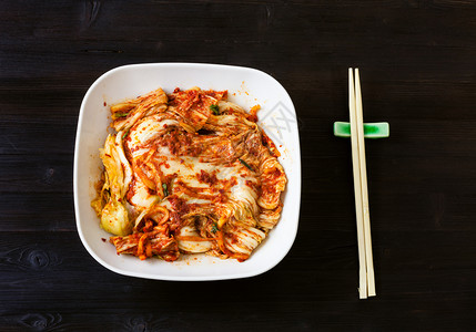 班禅korean烹饪白碗中Kimchi开胃菜辣椒小麦卷心菜和黑棕色木制餐桌上筷子的顶端视图背景