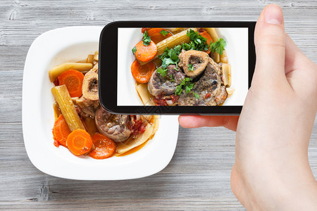 旅行概念智能手机用白色盘子上的意大利餐Osssobuco的游客照片背景图片