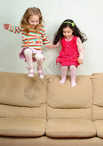 朋友们两个可爱的小女孩在沙发上跳跃两个小女孩在沙发上跳跃积极的运动背景图片