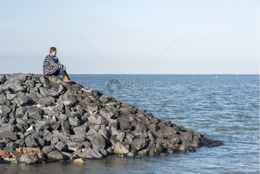 穿着棕靴子和蓝衣服的成年白人妇女坐在海边岩石上望着地平线等待某事和思考期待着坐蓝色的为了图片