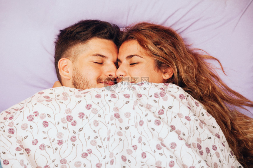 睡衣年轻笑着的异恋情侣躺在床上包裹枕头图片