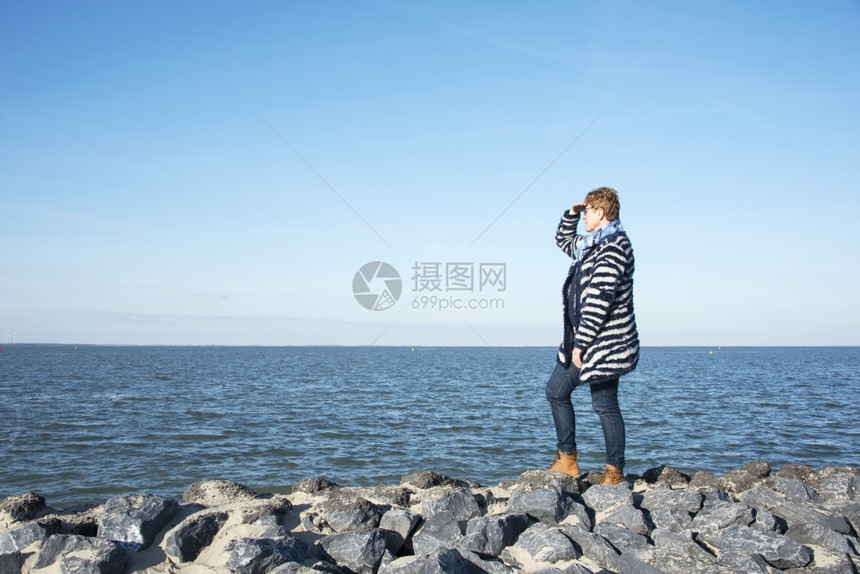 身着蓝衣服和棕靴子的成年妇女站在海岸的岩石上盯着空海的地平线看独自的棕色等待图片