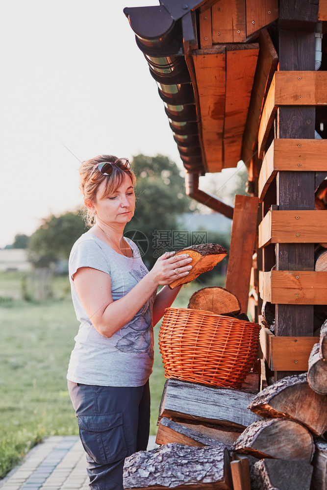 女士木制的柳条成年妇女把木柴原放在篮子上站后院的木棚旁真实人情况图片