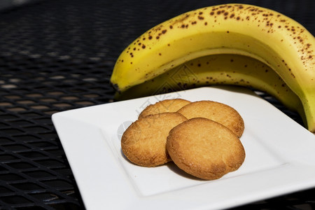 烘烤的正面美味奶油饼干和成熟的香蕉新鲜水果前白色盘子上的四块饼干在户外的黑金属庭院桌前健康背景图片
