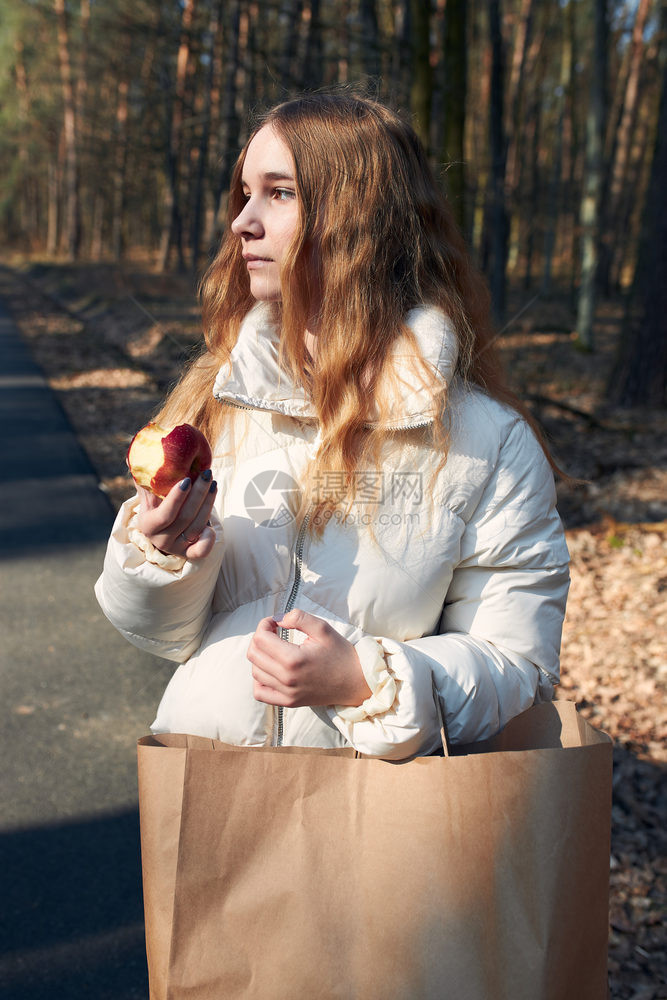 女孩站在公园看边拿着苹果穿白色夹克真实的人情况她穿着白色的夹克真实生活环境零售消费者纸图片