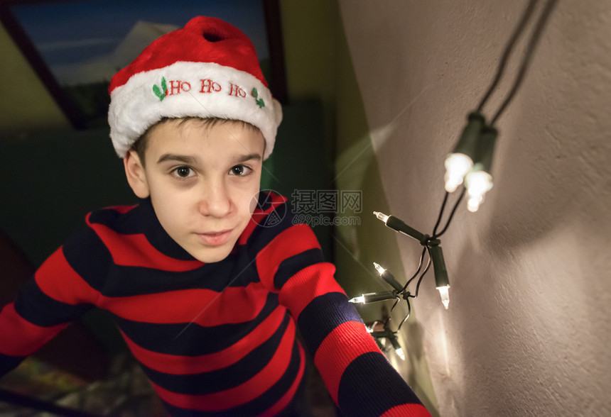 带着圣诞红帽子和灯光的男孩在他身边子穿着圣诞节服装看着摄影机自然真实的灯光孩子男生衣服乐趣图片