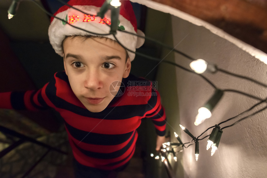 大约带着圣诞红帽子和灯光的男孩在他身边子穿着圣诞节服装看着摄影机自然真实的灯光孩子幸福真正的图片