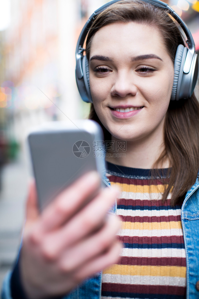 青年女子在街上走动通过移电话播放音乐城市年轻的耳机图片