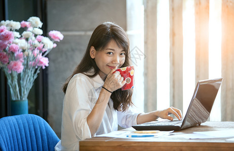 年轻亚洲女在自由职业者工作时喝着热咖啡的快乐笑脸手表眼睛更年轻图片