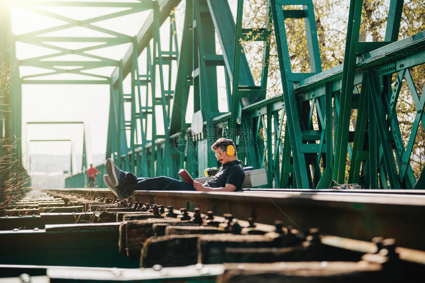 年轻笑的建筑工人坐在读一本关于旧钢铁路桥的书火车老木制图片