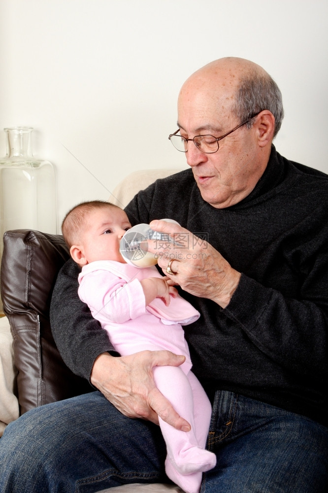 他的爸祖父用一瓶牛奶喂可爱的女婴同时坐在沙发上一个手抱着婴儿的起居室沙发上图片
