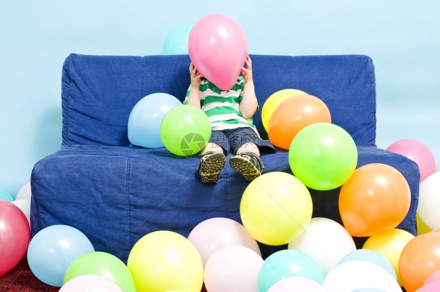 年轻男孩坐在沙发上躲棕榈树后面覆盖气球包围图片