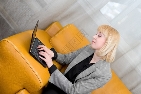 金发年轻女青用笔记本电脑坐在皮沙发上金女郎黄色的支持图片