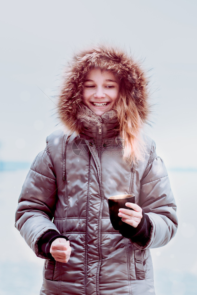 一种笑的年轻女孩在冬天散步时喝热饮穿着戴头罩的外套拿着茶杯喝美丽走图片