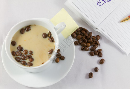 早晨咖啡店一杯加牛奶的咖啡一本开放式笔记白底带一枝笔和咖啡豆黑色的图片