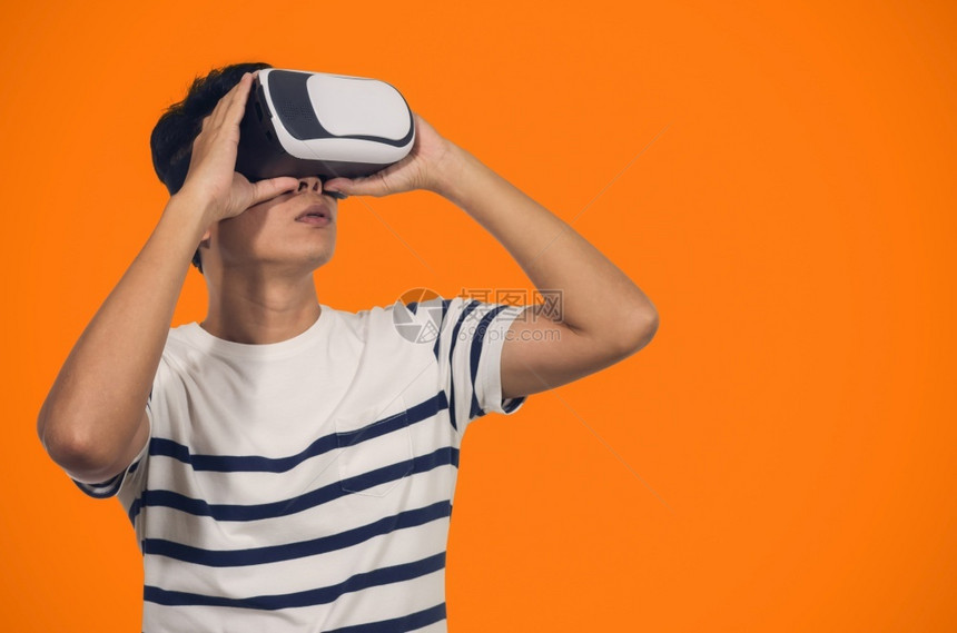 接触面具有虚拟现实头盔的年轻人站在橙色背景的室内青年拿着虚拟现实头盔的年轻人眼镜图片
