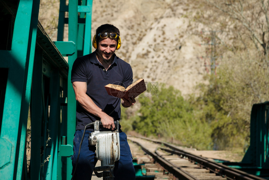 火车年轻建筑工人在读一本关于旧钢铁路桥的书时手里拿着液压锤子木制的听到图片