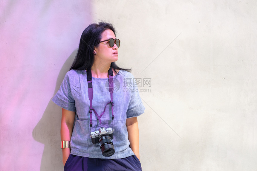 摄影机靠在混凝土墙上日光时装旅行者身穿太阳镜的高级亚洲人相机图片