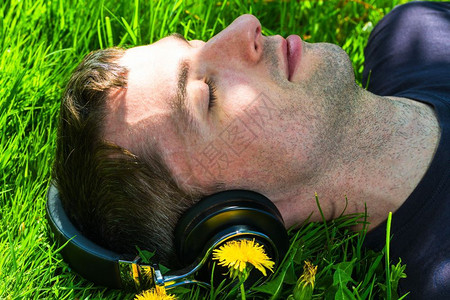 耐用的全尺寸新青年男子用无线耳机监听音乐躺在草坪上与dandelions背景图片