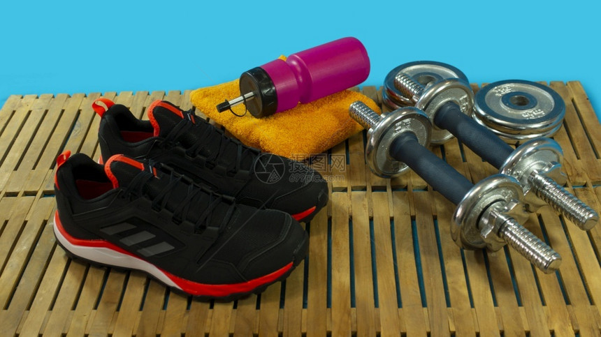 健身房一套黑色运动鞋用哑铃黄色毛巾和蓝背景木地板上带水的红色瓶子运动木制钳图片