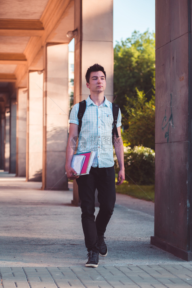男学生拿着笔记本背着书包走在大学楼的走廊里图片