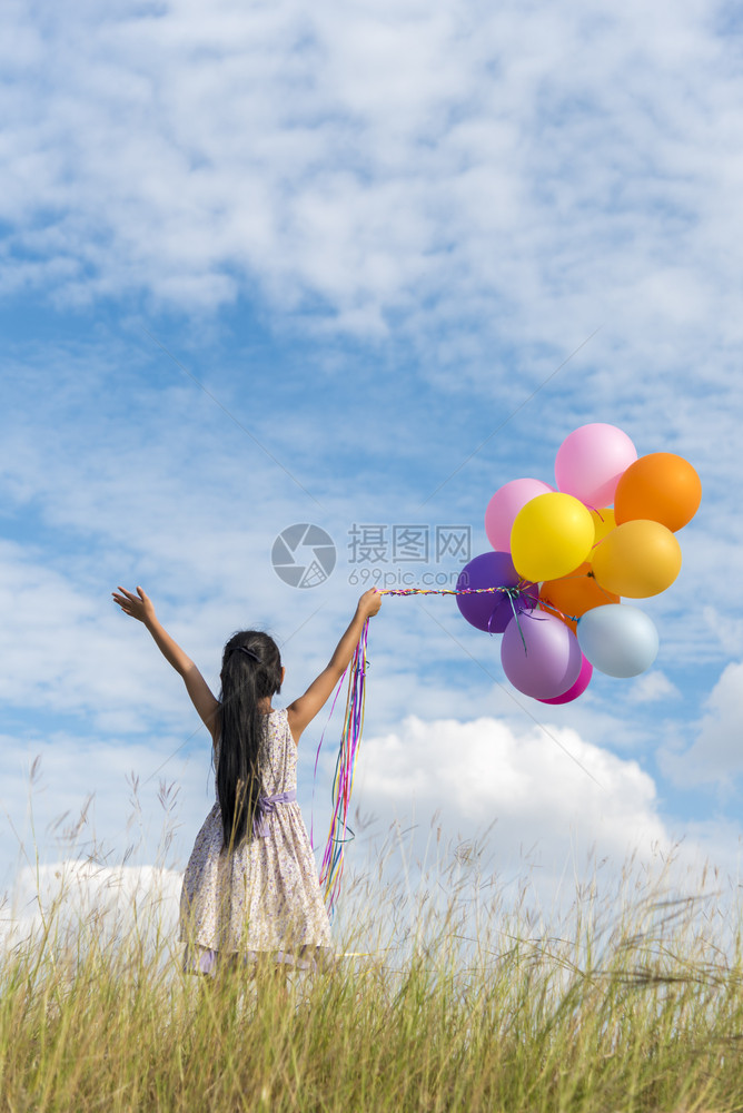 快乐的女孩在一片充满云彩蓝天的绿草地上拿着多彩空气球夏天孩子们户外图片