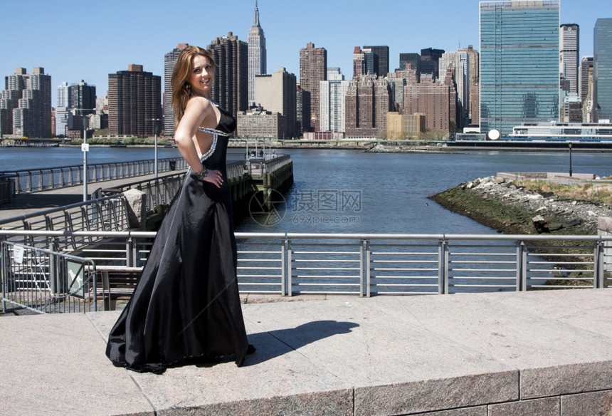 晚上穿着黑鸡尾酒装扮的美丽笑着快乐高加索拉丁时装模特女郎站在纽约市曼哈顿天际线前联合的新图片
